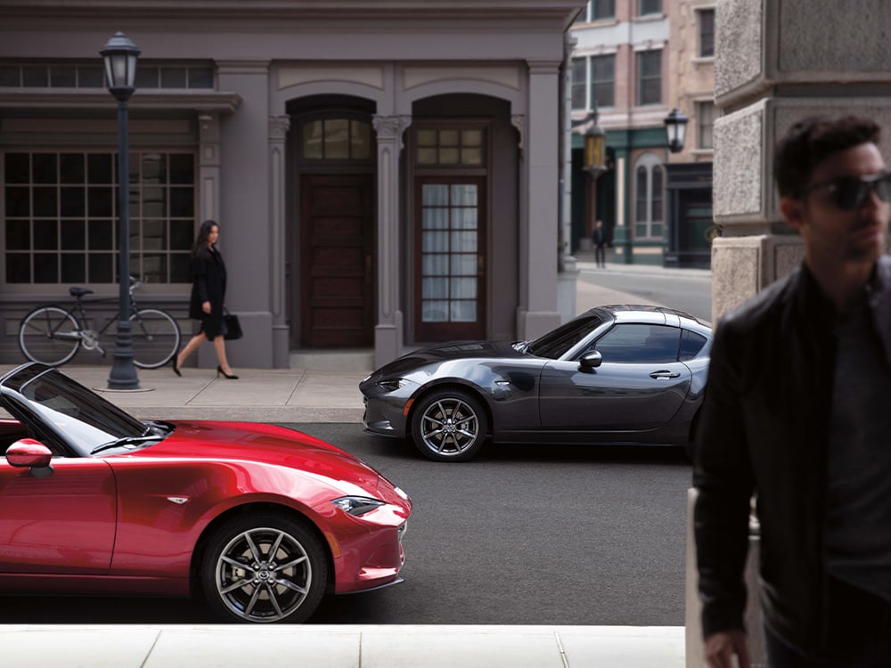 Two gleaming Mazda MX-5s parked along urban street, a woman looks in the background as a man walks away in the foreground.
