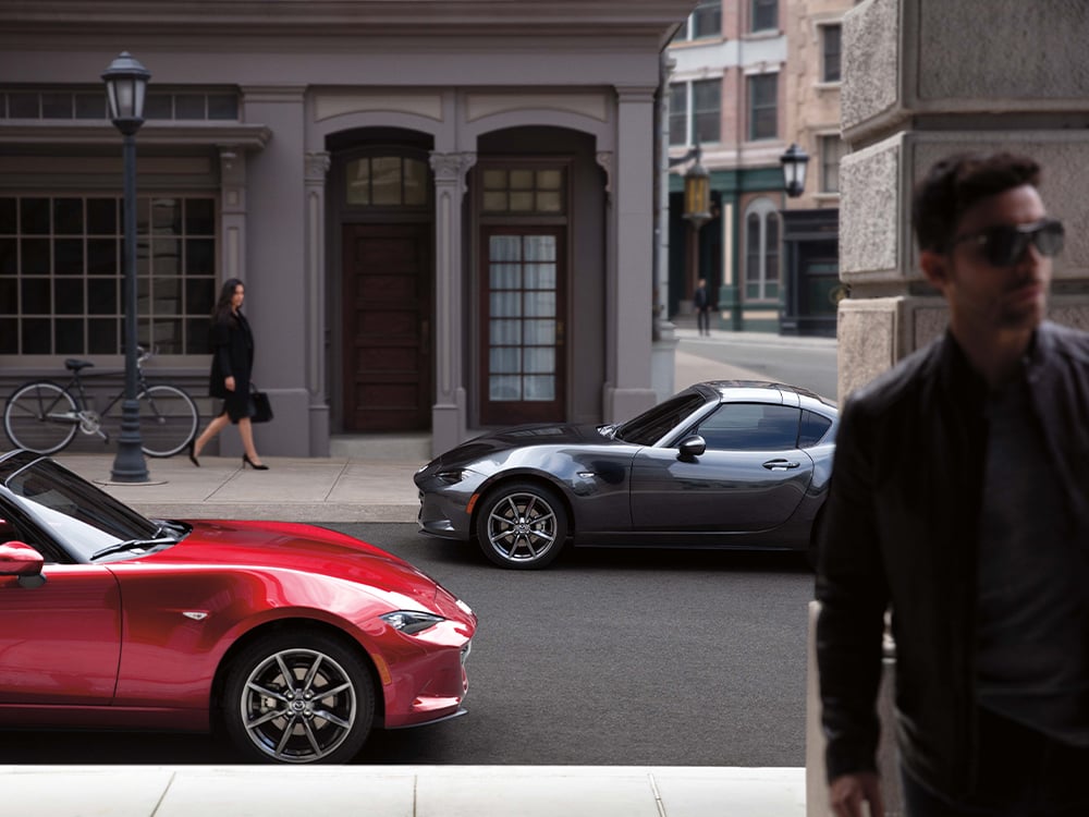 Deux rutilantes Mazda MX-5 garées le long d’une rue urbaine; une femme regarde en arrière-plan tandis qu’un homme s’éloigne au premier plan.