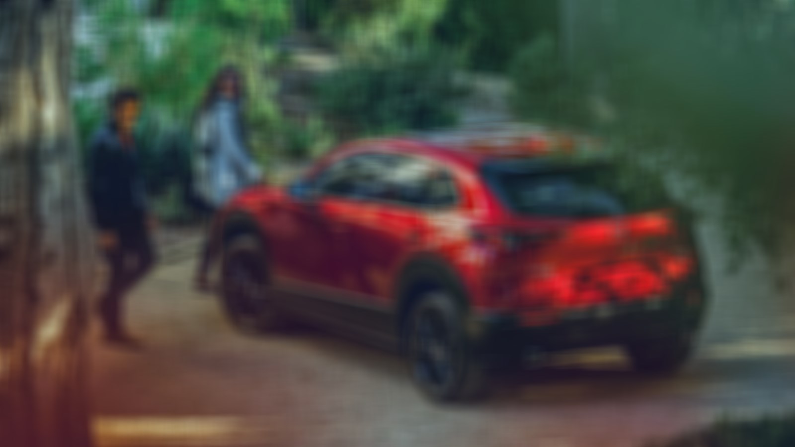 Couple approaches Soul Red Crystal Metallic CX-30 parked in the shade before a rustic trail entrance. 