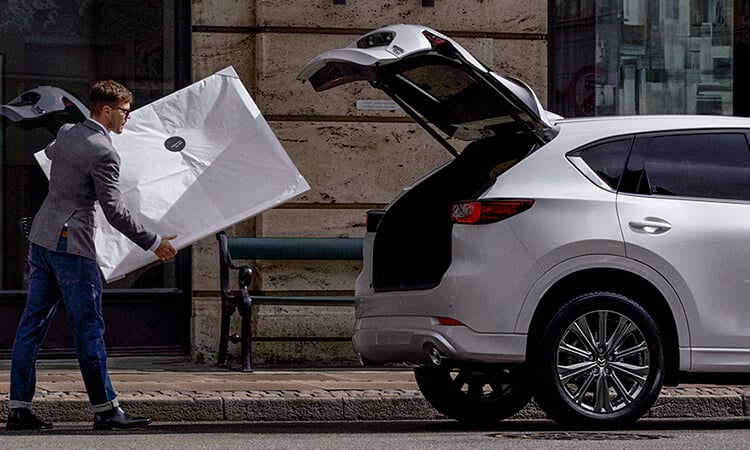 Man carries white frame to Rhodium White Metallic Mazda CX-5 parked on city street with liftgate open. 