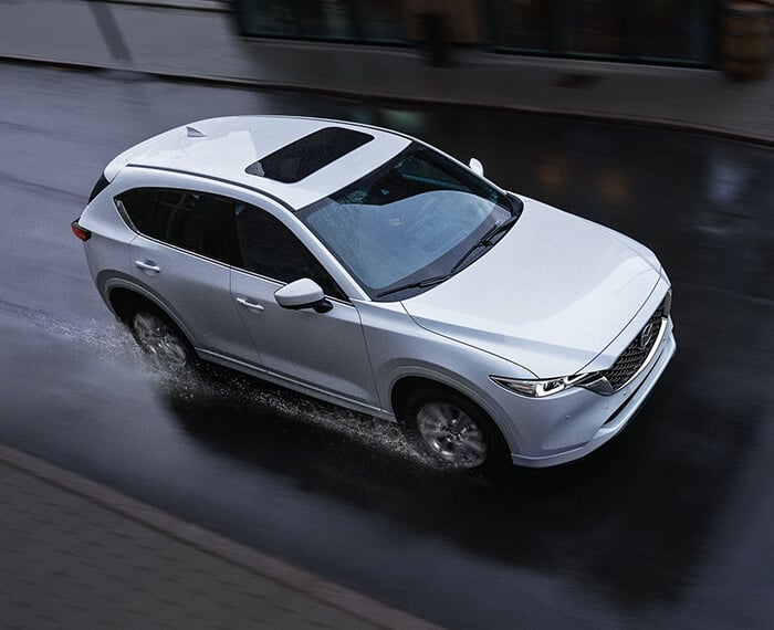Rhodium White Metallic Mazda CX-5 kicking up water, driving down rain-soaked city street.