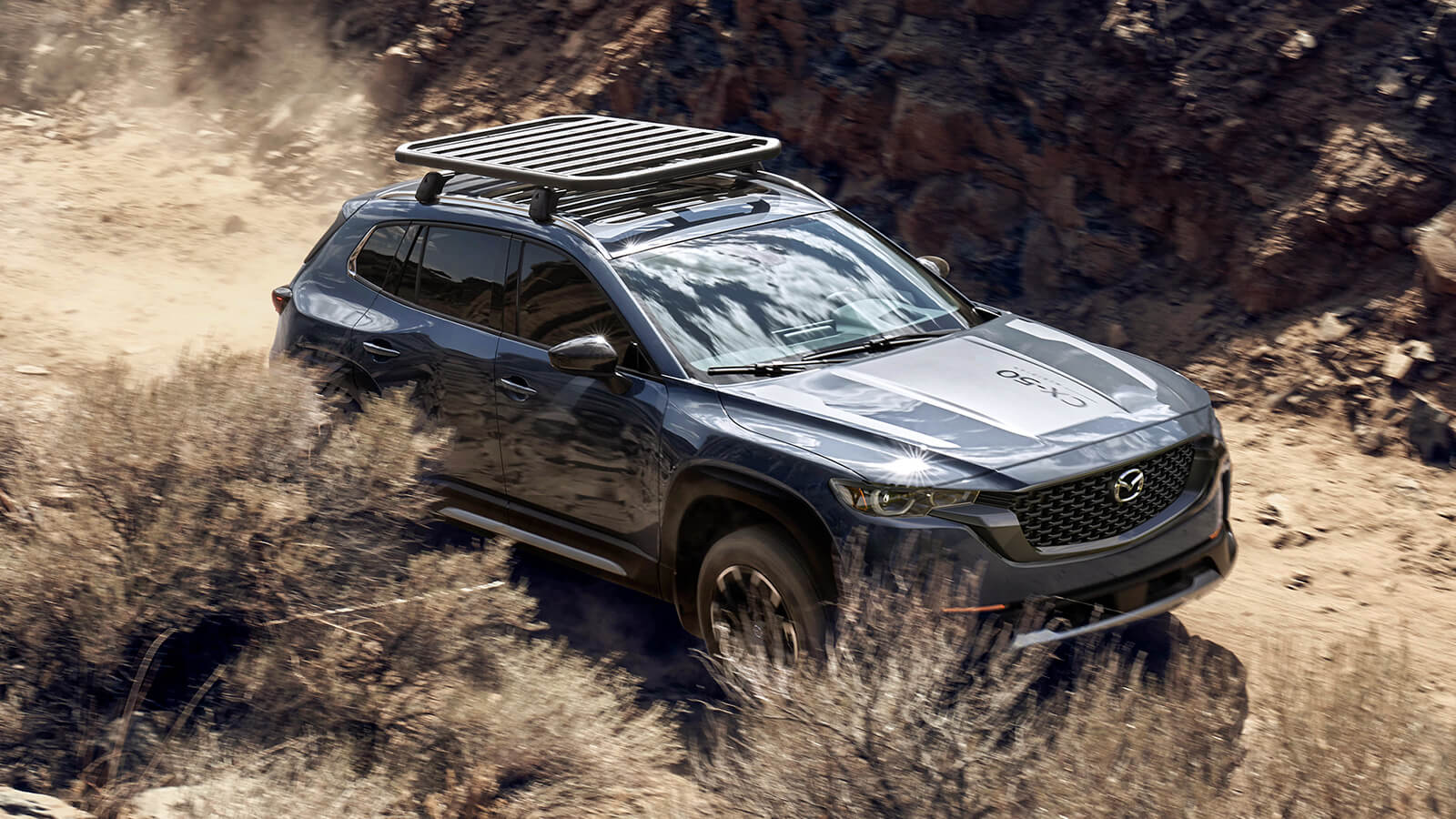 CX-50 with roof rack drives along dusty path. 