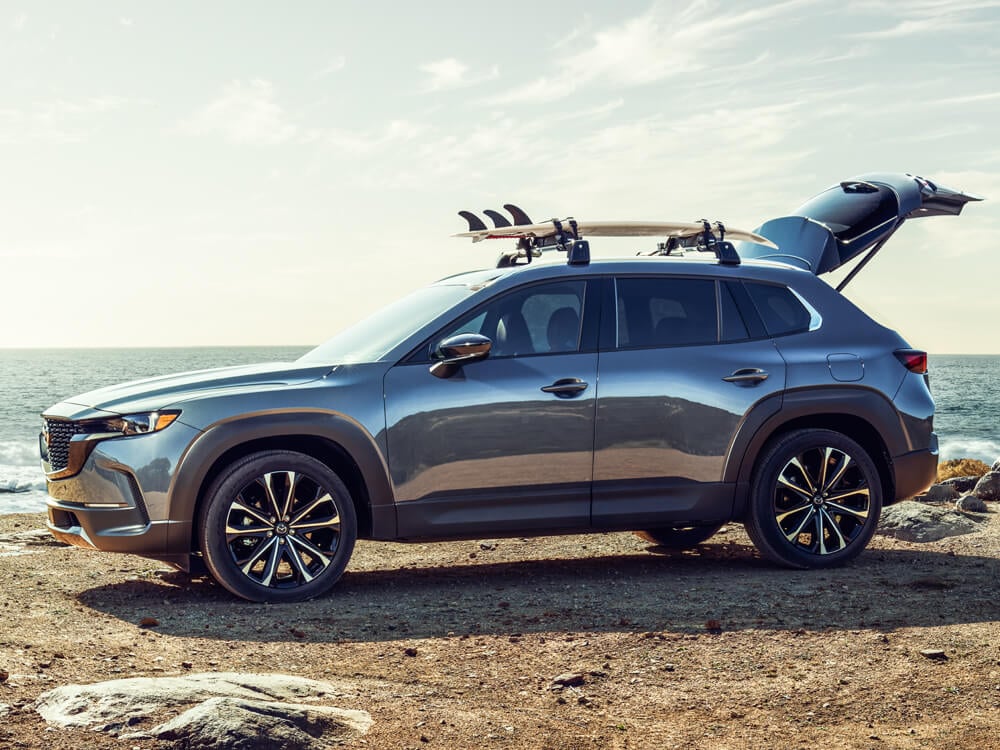 Polymetal Grey Metallic CX-50 parked on beach with back liftgate ajar, a surfboard is stored on the roof rack. 