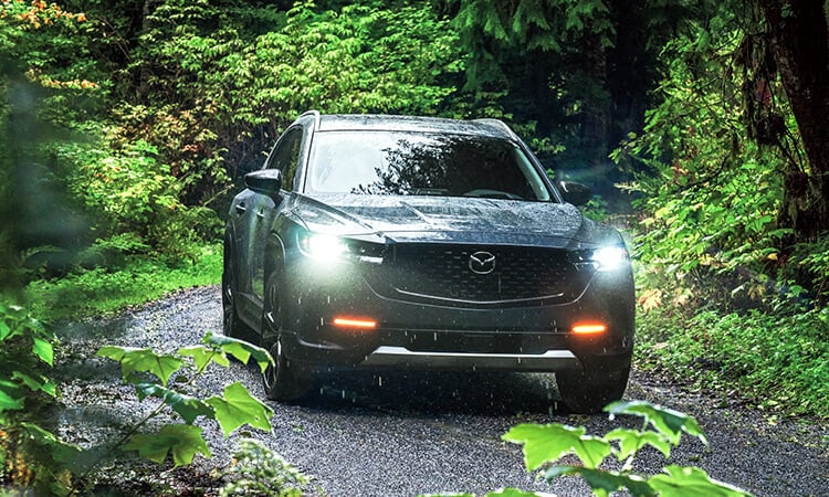 Ingot Blue Metallic CX-50 with headlights on, driving in dense forest. 