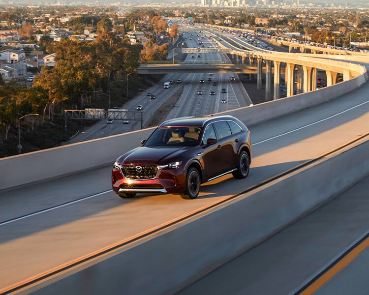 Artisan Red CX-90 PHEV drives toward sunset on concrete highway overpass.