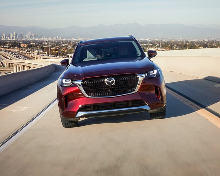 Front shot of Artisan Red CX-90 PHEV driving towards camera casting a shadow on highway overpass.