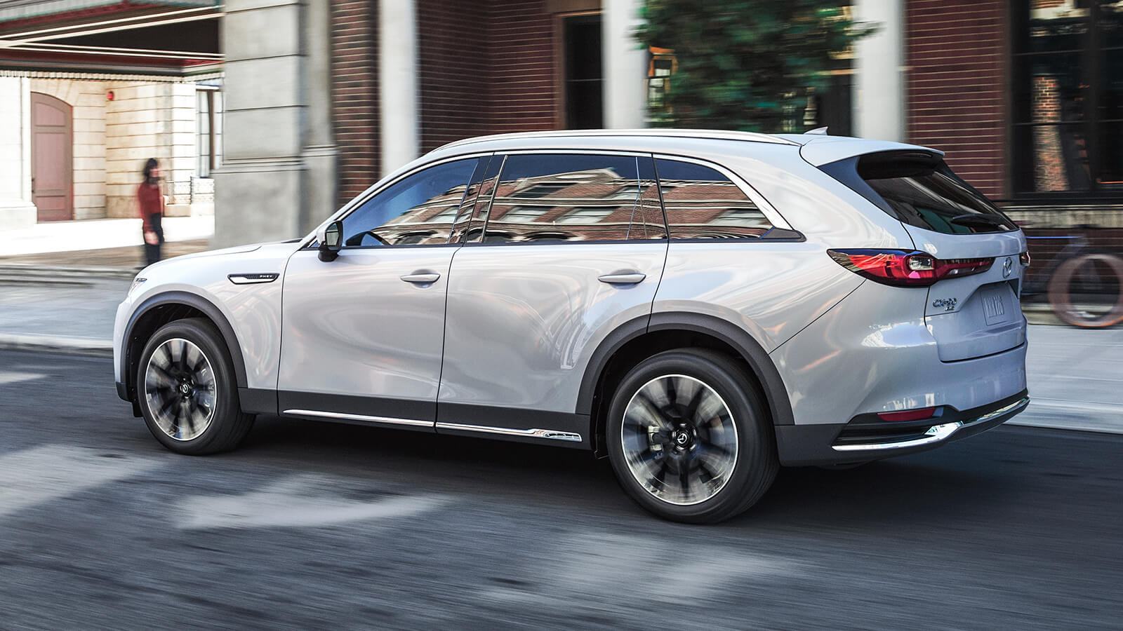 Artic White CX-90 PHEV passes a pedestrian in front of red brick building on city street