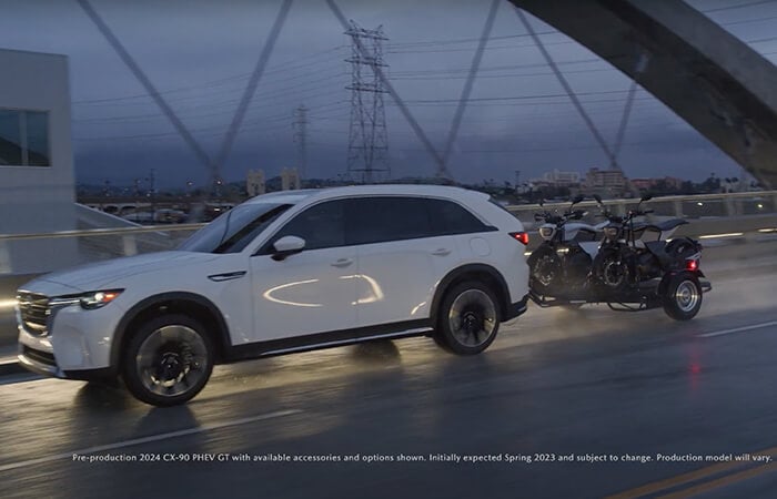 A CX-90 PHEV driving down a wet road while towing two motorcycles.