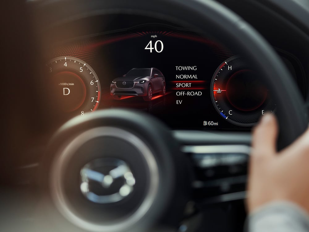 The CX-90 dash displays driver toggling through Mi-Drive modes through the steering wheel, selecting Sport. 
