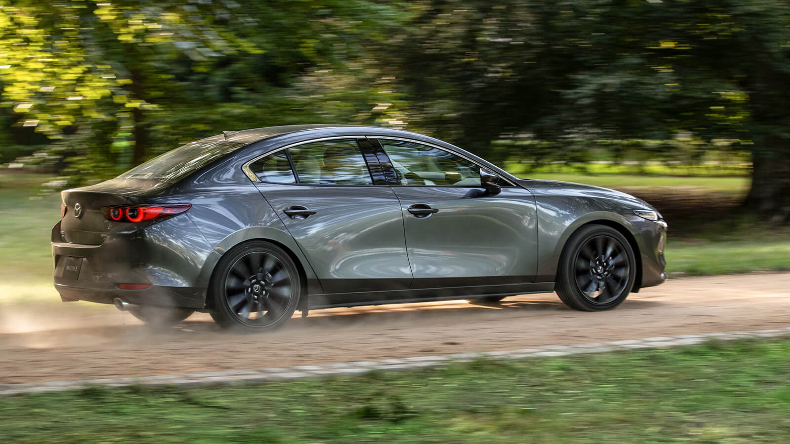 Mazda3 sedan drives swiftly along country road kicking up dust, passing a large tree in the background. 