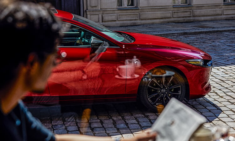 Un homme attablé sur la terrasse d’un café regarde son reflet dans une Mazda3 Sport rouge vibrant cristal métallisé garée dans une rue pavée. 