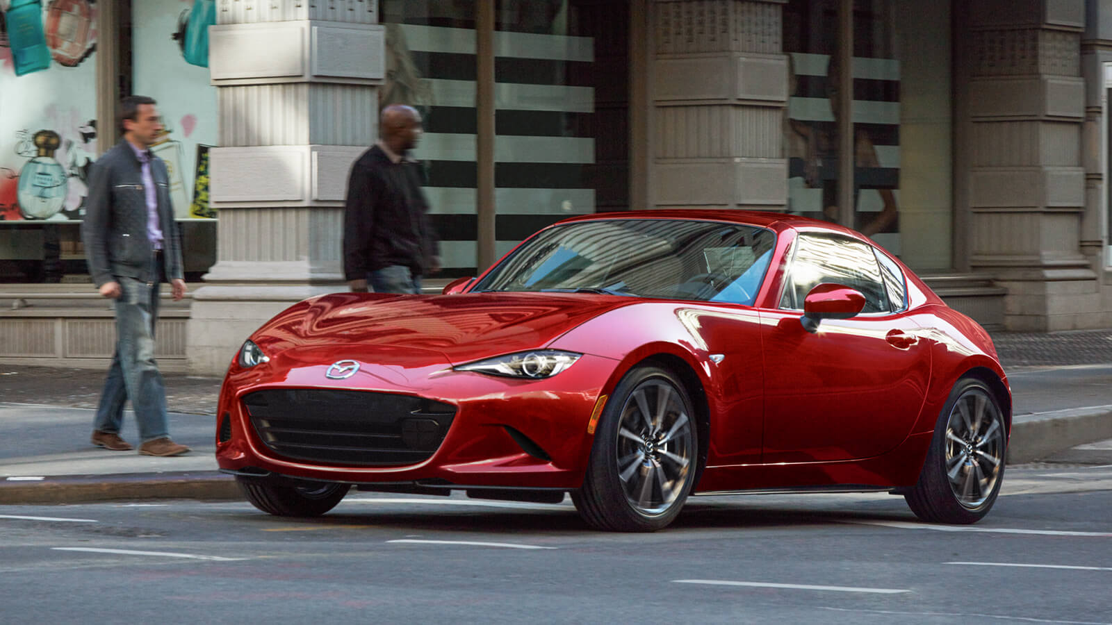 Soul Red Crystal Metallic MX-5 RF with the top up pulls away from city crosswalk, pedestrians wait to cross on the corner in the background.