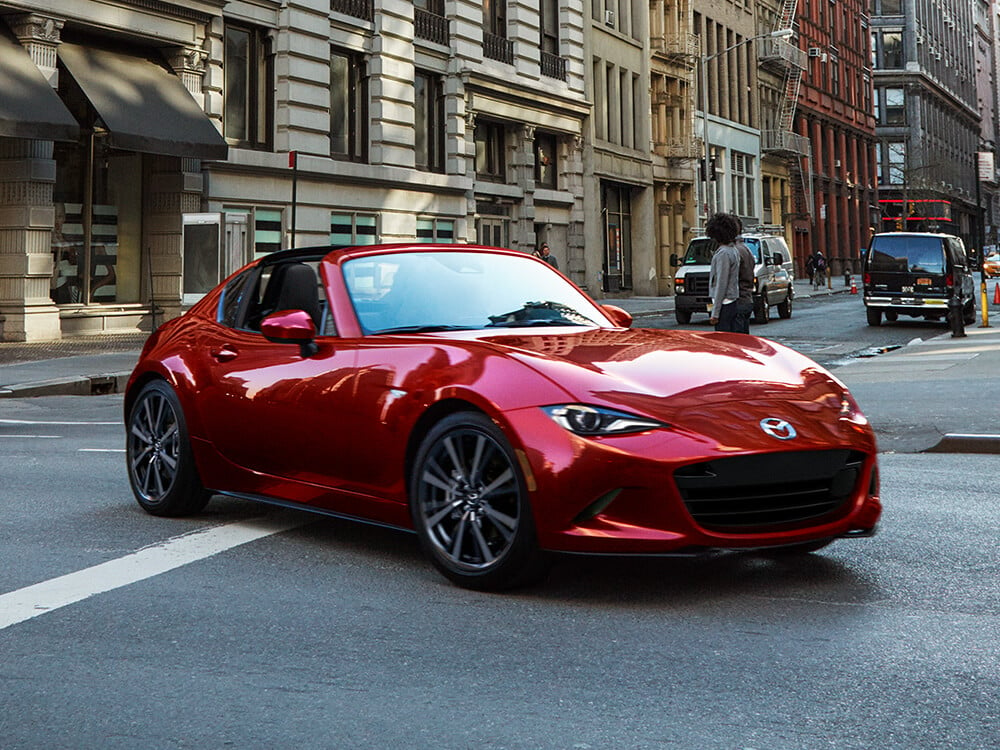 Soul Red Crystal Metallic MX-5 RF reflects the surrounding buildings as it turns left onto sideroad in a busy city. 