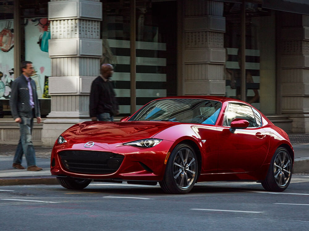 Soul Red Crystal Metallic MX-5 RF with the top up pulls away from city crosswalk, pedestrians wait to cross on the corner in the background. 