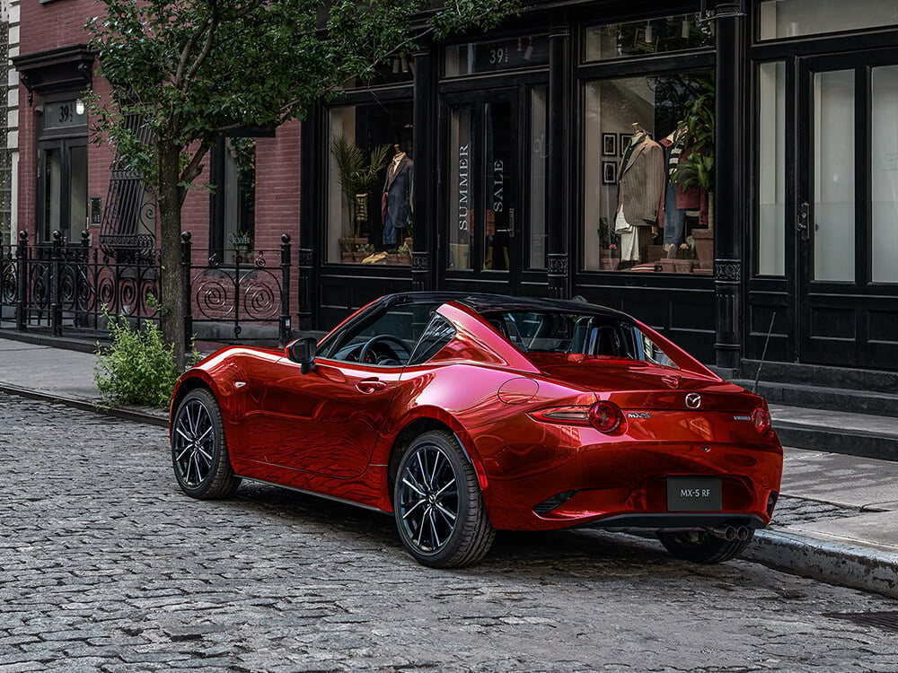 A gleaming Soul Red Crystal Metallic MX-5 RF with the top down is parked along urban cobblestone street, in front of formal menswear shop. 