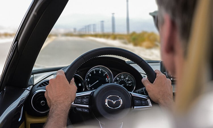Shot from behind the driver inside the MX-5 ST, hands on the wheel, he looks out to an open road lined with utility poles. 