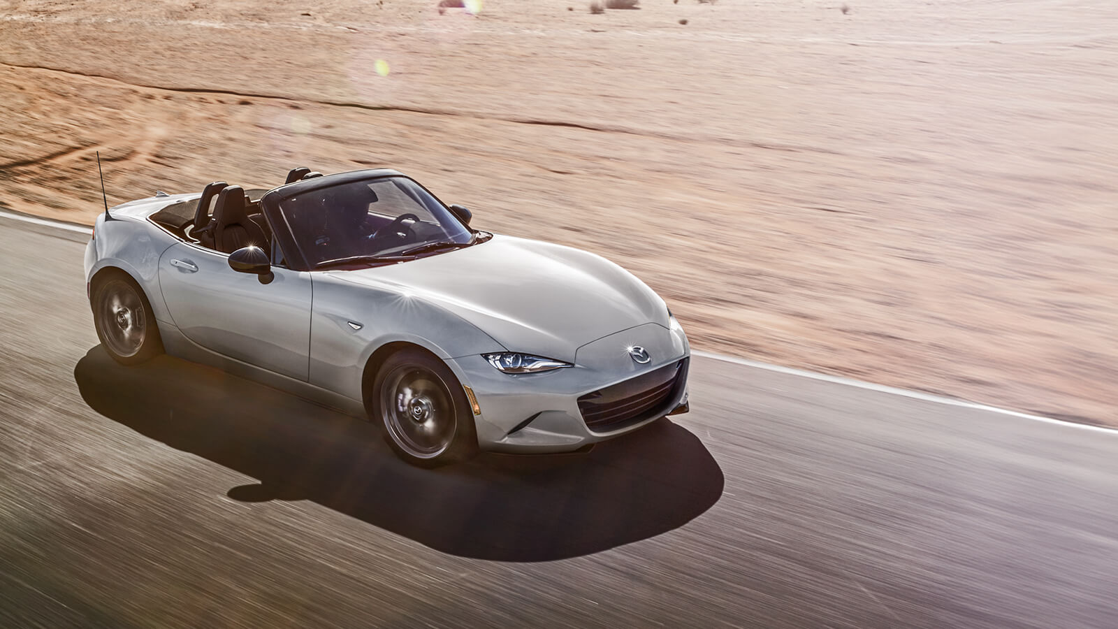 A lustrous Snowflake White Pearl MX-5 ST reflects sunlight as it drives down paved road next to sandy beach. 