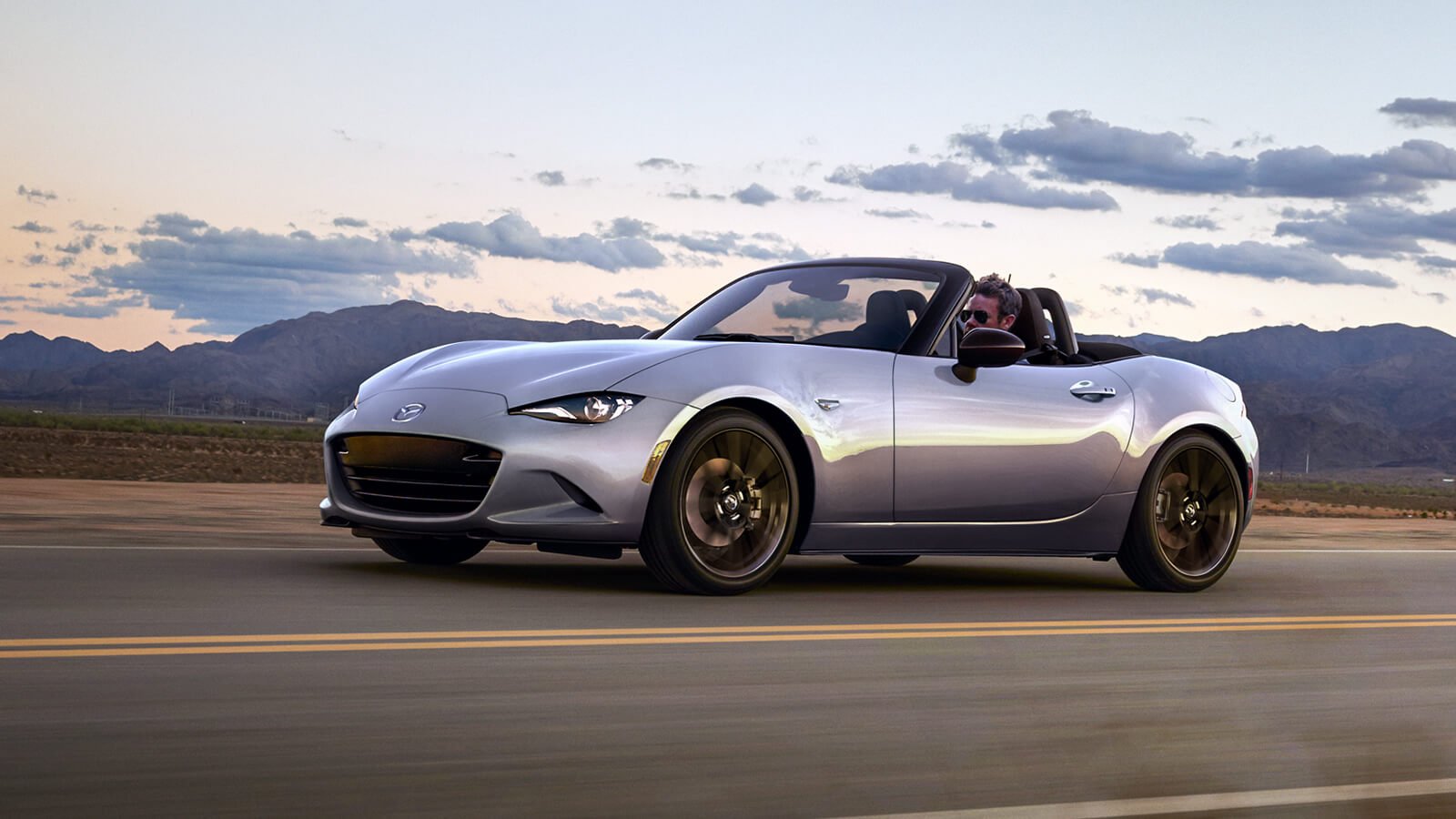Machine Grey Metallic Mazda MX-5 Soft Top with roof open rounds drives down highway on a blue-sky evening beyond a mountain range. 