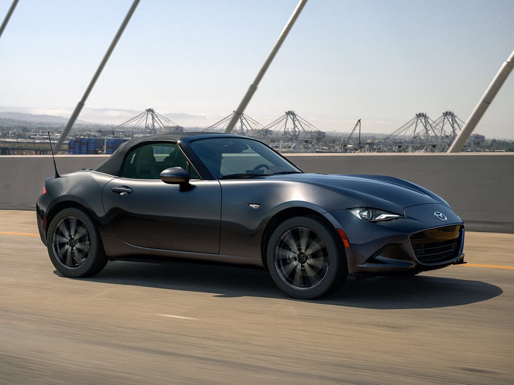 Machine Grey Metallic MX-5 ST with the top up drives over service bridge with an industrious city background.