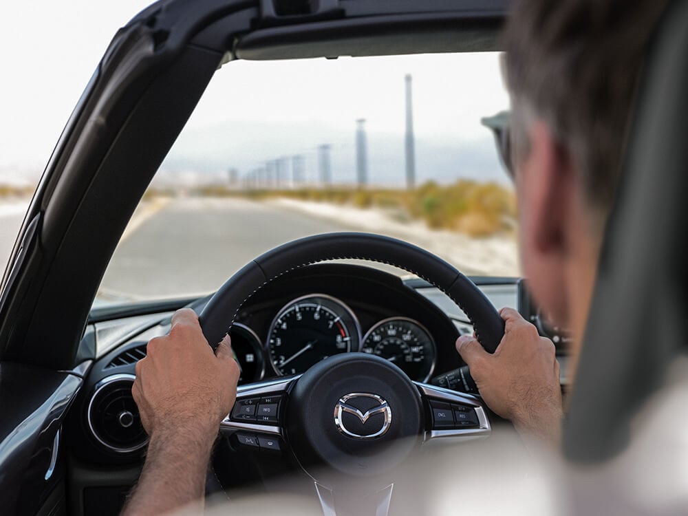 Shot from behind the driver inside the MX-5 ST, hands on the wheel, he looks out to an open road lined with utility poles.