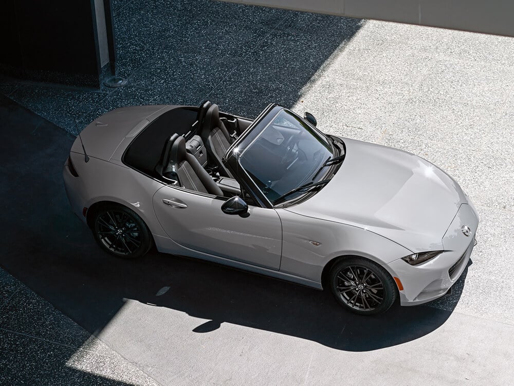 Top down shot from the passenger’s side of an Aero Grey Metallic MX-5 ST with the top down, the back is parked in a shadow cast by the surrounding building while the front shines in midday sun.  