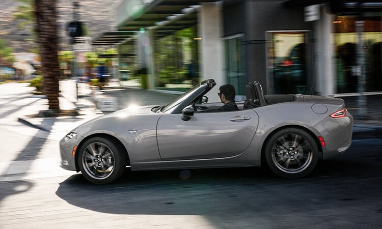 Driver’s side profile view of Machine Grey Metallic MX-5 turning right on city street. 
