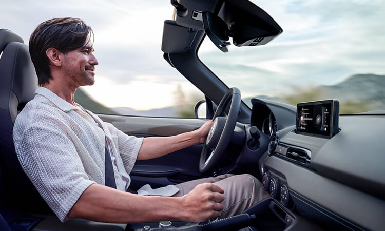 Vue d’un conducteur souriant avec sa main sur le pommeau de levier de vitesses depuis le siège passager à l’intérieur d’une MX-5. 