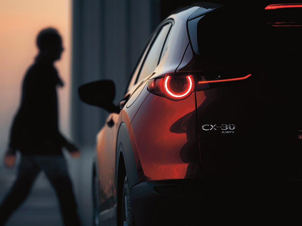 Close up of rear driver’s side of Soul Red Crystal Metallic CX-30 parked at dusk, focusing on CX-30 and AWD badge. Red tailight is illuminated. In the background, an out of focus silhouette of a man walking.