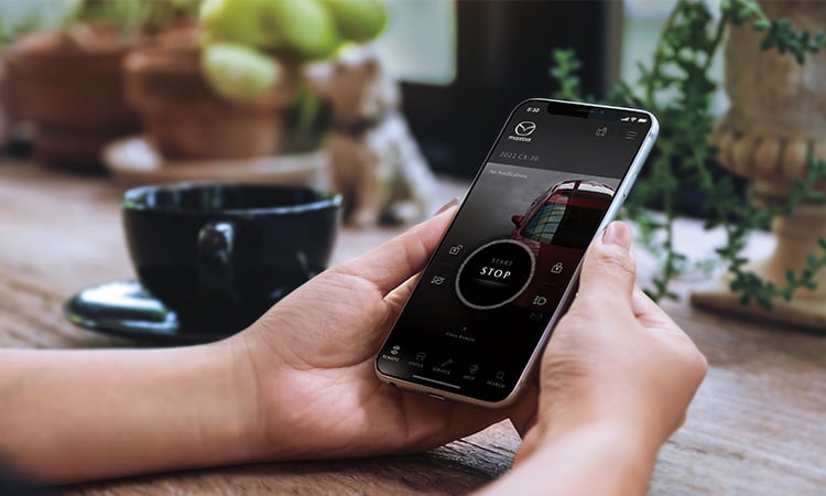 Hands cradle smartphone displaying MyMazda app start screen.