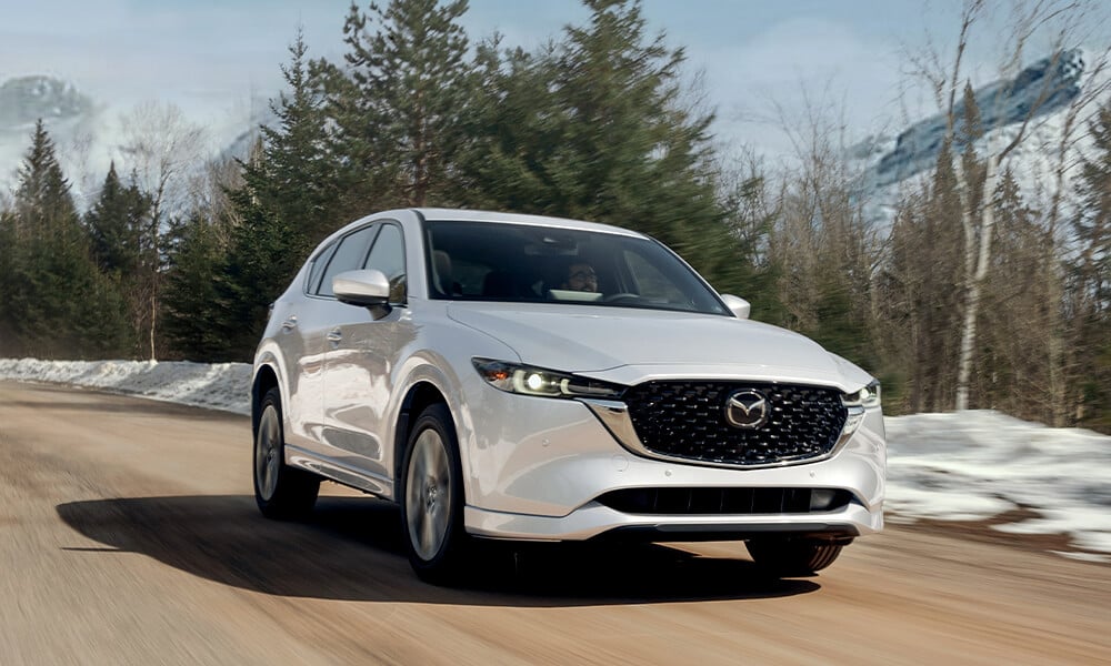 Rhodium White Metallic Mazda CX-5 ¾ front view driving on paved road with snowy blurred background.