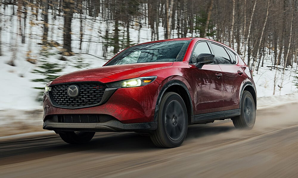 Soul Red Crystal Mazda CX-5 driving, side-front view, with snowy forest blurred in background.