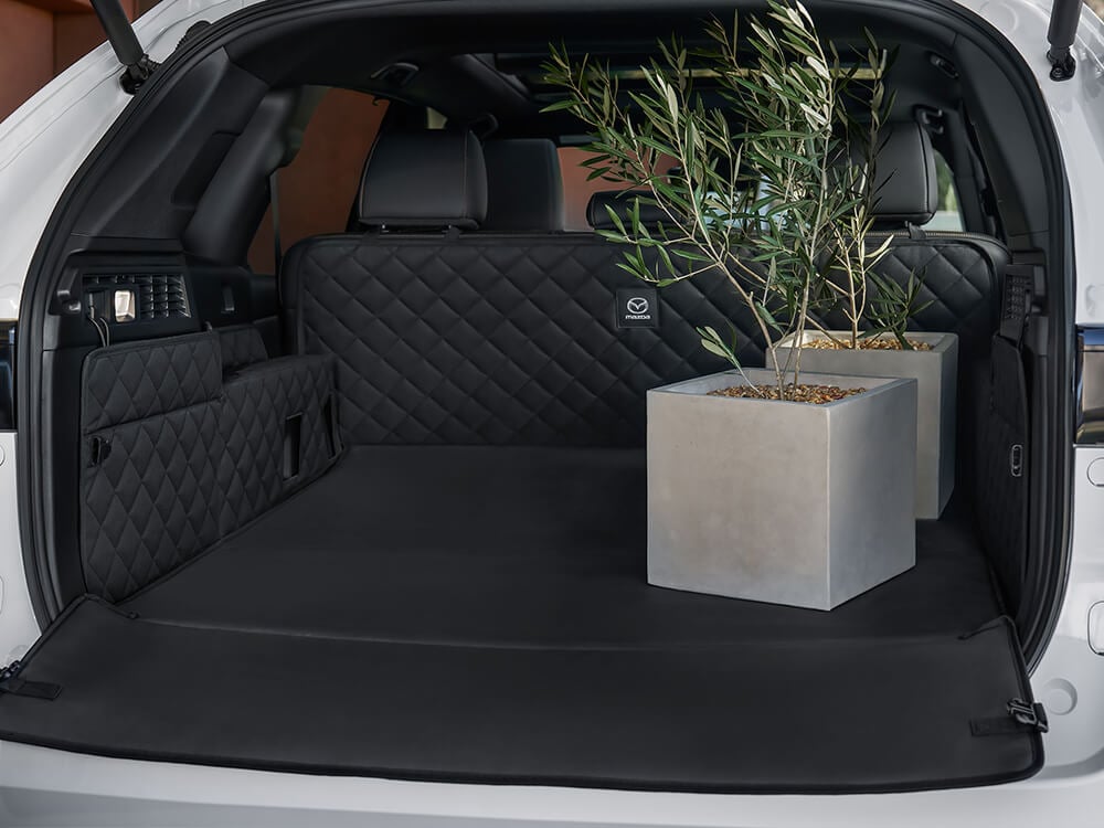 Shot from behind the CX-70 PHEV, an open liftgate reveals the expansive storage space with quilted protectors and two large potted plants inside.