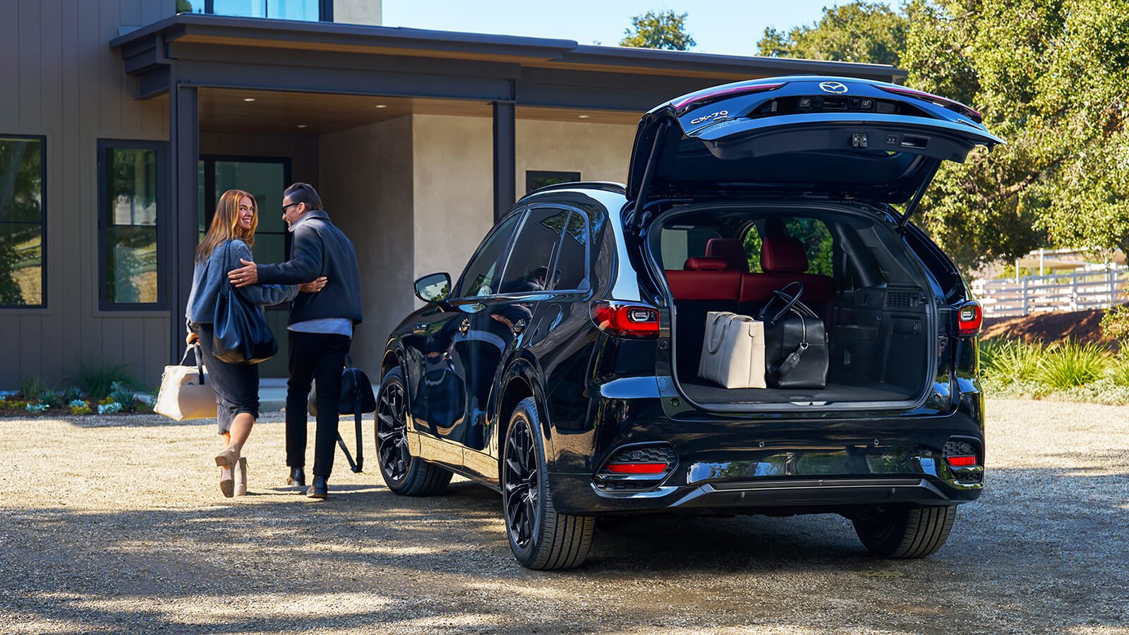 Parked CX-70 reflects the surrounding driveway at verdant modern home, view from the back with lift gate open revealing luggage as a smiling couple approaches the home.