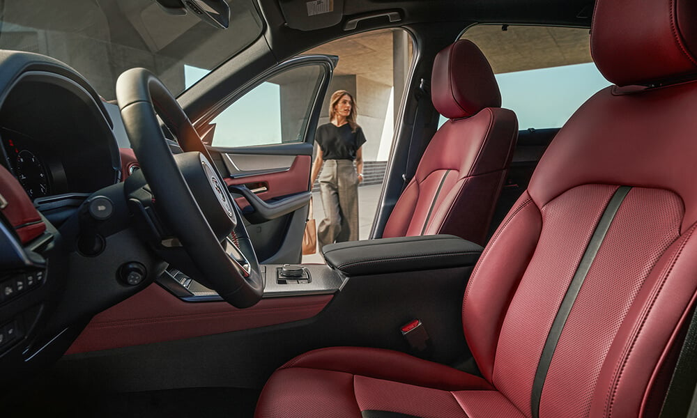 Shot from driver’s side inside the CX-70, Burgundy Nappa leather seats, a passenger walks toward the open passenger side door.