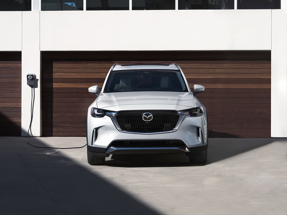 Un CX-90 hybride rechargeable blanc branché à une borne domestique dans l’allée d’une maison avec une porte de garage en bois.