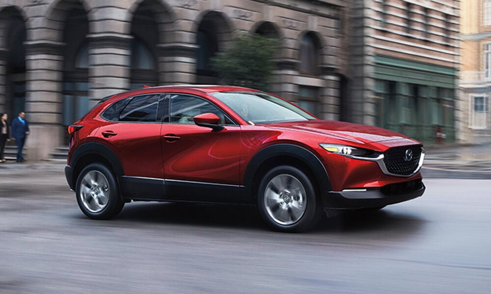 Vue de profil d’un Mazda CX-30 rouge vibrant cristal métallisé roulant en ville avec des bâtiments et des piétons flous pour transmettre un effet de mouvement.