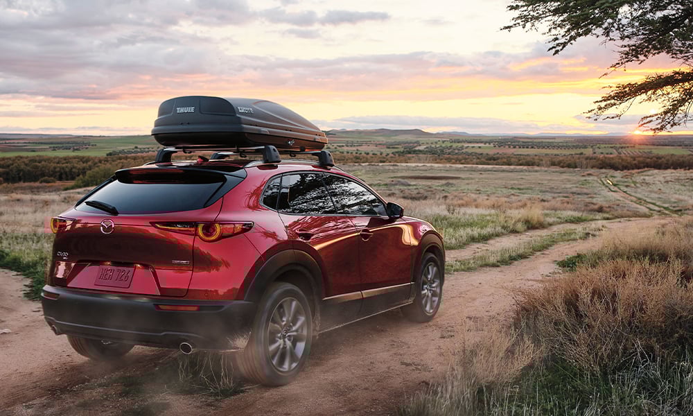 n homme et une femme se tiennent près du coffre d’un Mazda CX-30 rouge vibrant cristal métallisé avec un paysage extérieur flou en arrière-plan.