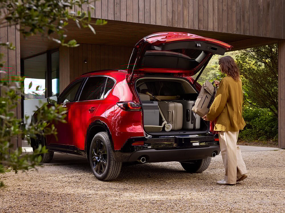 Profil latéral arrière de trois-quarts d'un Mazda CX-5 rouge vibrant cristal avec le coffre ouvert, alors qu'une femme place la dernière valise dans un coffre déjà rempli de bagages dans son entrée.