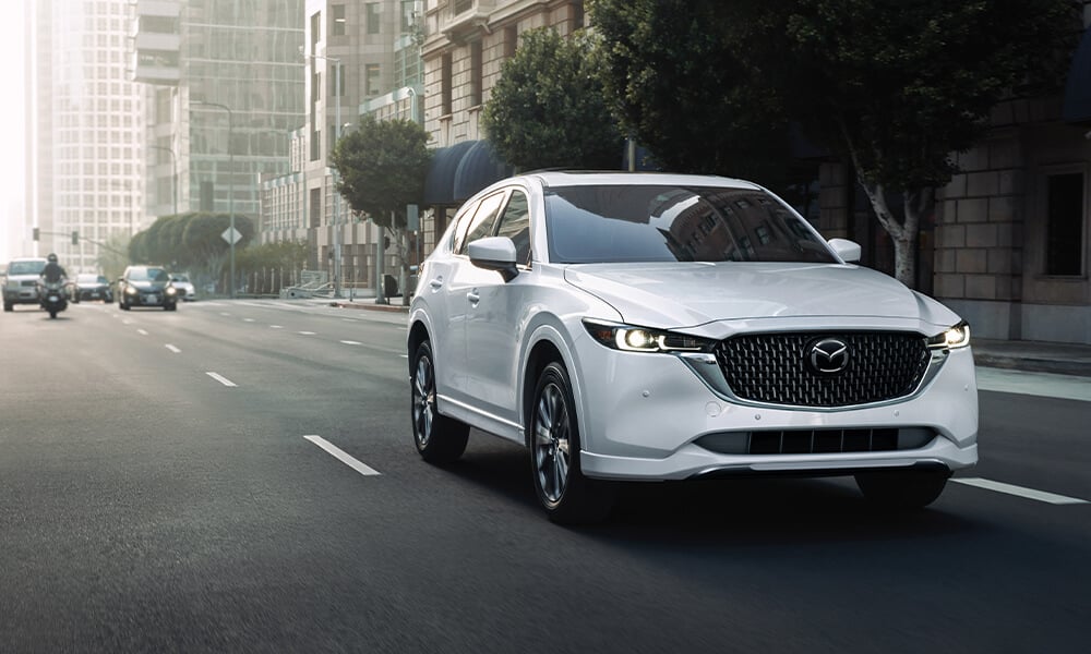 Profil latéral avant d'un Mazda CX-5 blanc rhodium métallisé roulant vers la caméra sur une route urbaine.