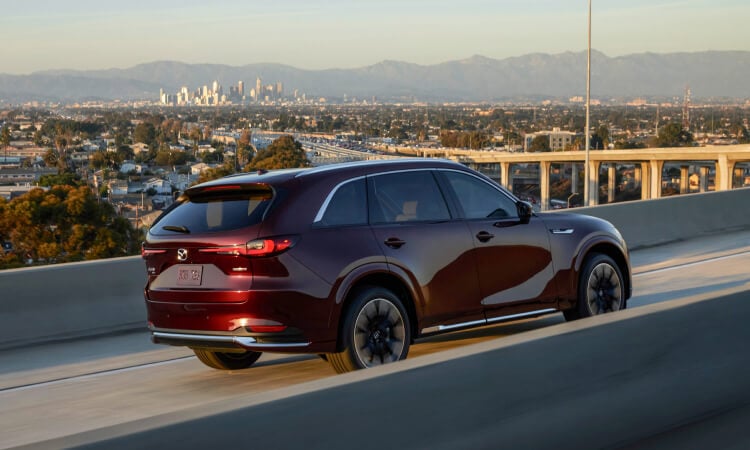 Side profile of a CX-90 driving on a highway with a city view in the background.