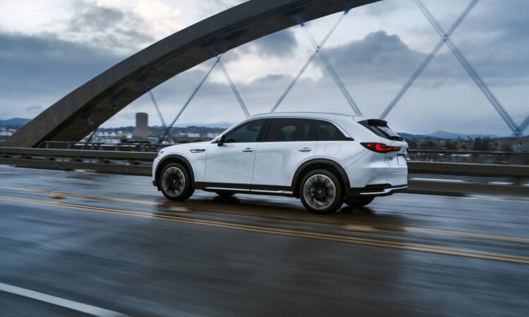 Side profile of the CX-90 driving across a rain-soaked bridge.