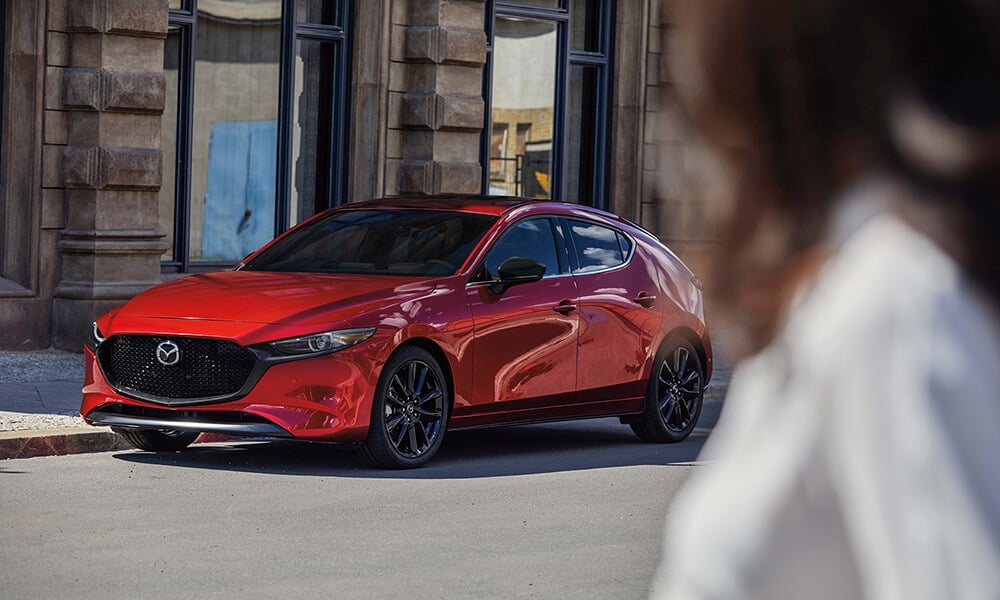 Une femme floue en avant-plan regardant vers une Mazda3 Sport rouge vibrant cristal métallisé montrée de profil.