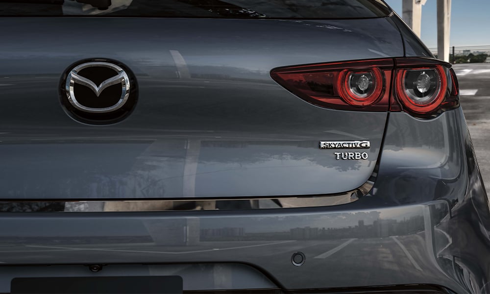 Vue de l’intérieur d’une Mazda3 Sport à travers le toit ouvrant panoramique, montrant une main posée sur le bouton de commande central.