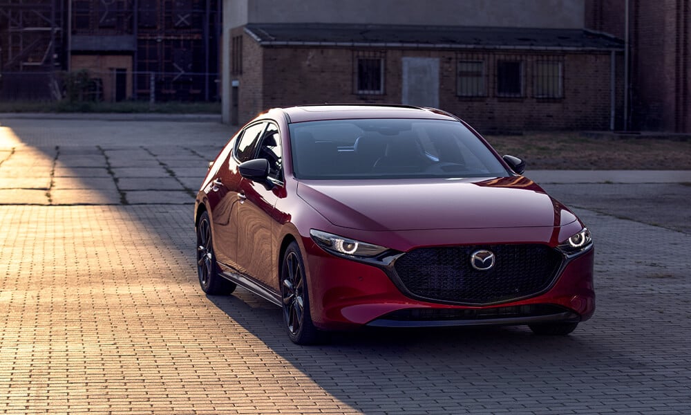 Vue avant d’une Mazda3 Sport rouge vibrant cristal métallisé stationnée dans l’ombre d’un immeuble illuminé par le soleil.