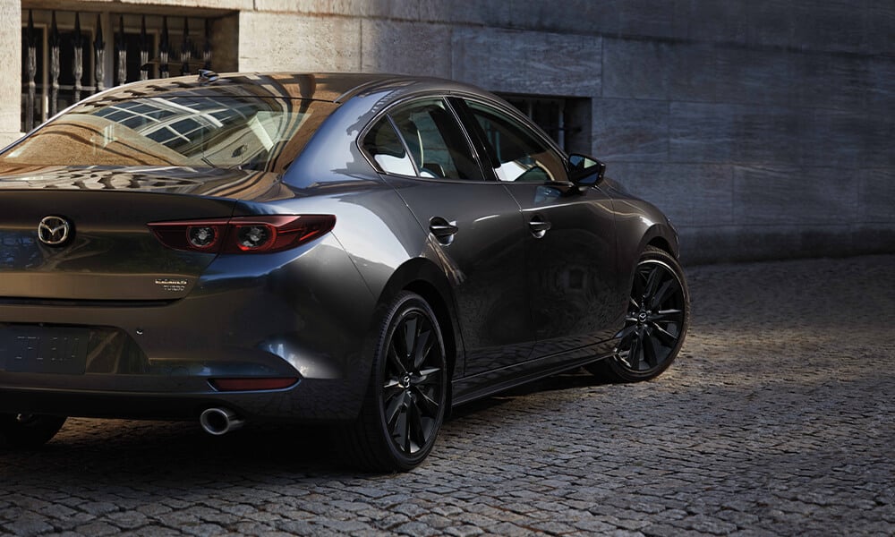 Back view of a Machine Grey Metallic Mazda3 with the front wheel turned, driving on a cobblestone road.
