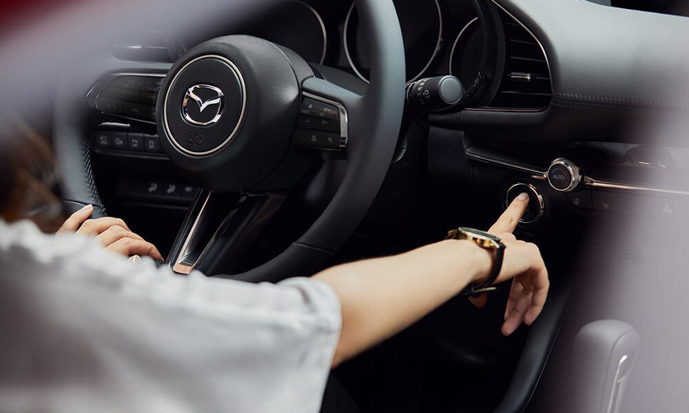 Over-the-shoulder view of a woman in the Mazda3 pressing the engine start button with one hand and holding the steering wheel with the other. 