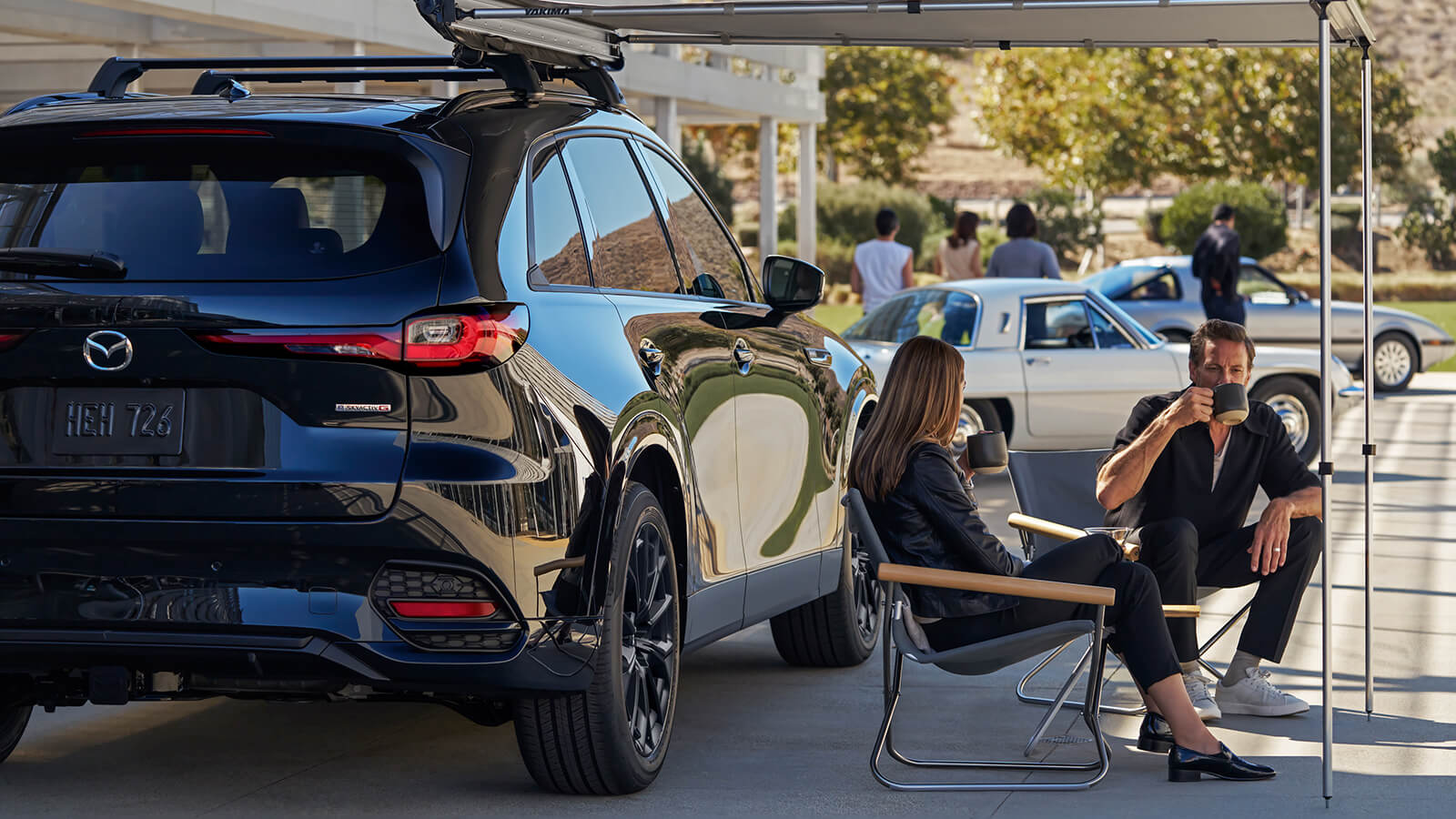 Woman sits with partner sipping from a coffee mug under a pop-up tent at a parking lot car-meet with their Jet Black Mica CX-70.